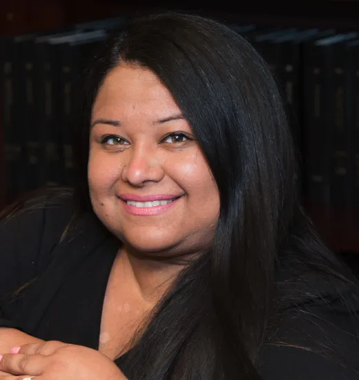 A woman with long black hair sits thoughtfully in front of a bookcase filled with books.
