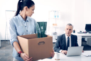 Selective focus on a serious chief working on a laptop and looking at his former office worker leaving the office with a box full of different supplies.