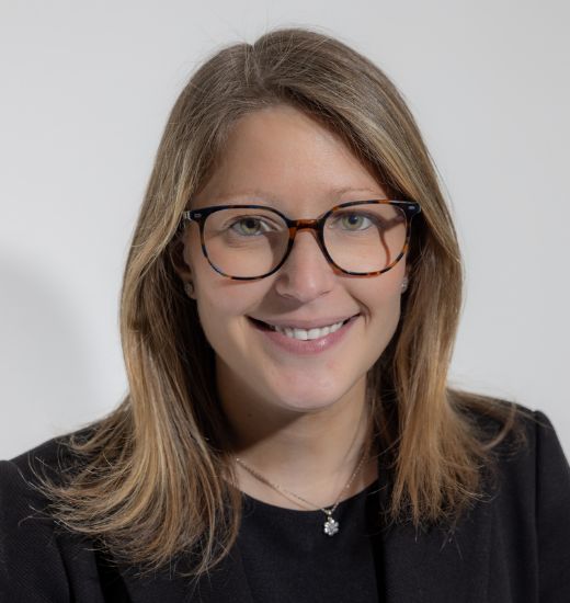 A woman wearing glasses and a black blazer smiles warmly, exuding confidence and professionalism.